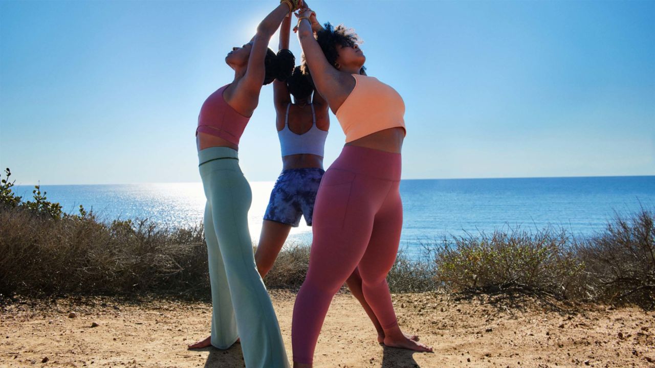 Three people wearing Athleta activewear are facing away from each other form a circle with their hands up in the air.