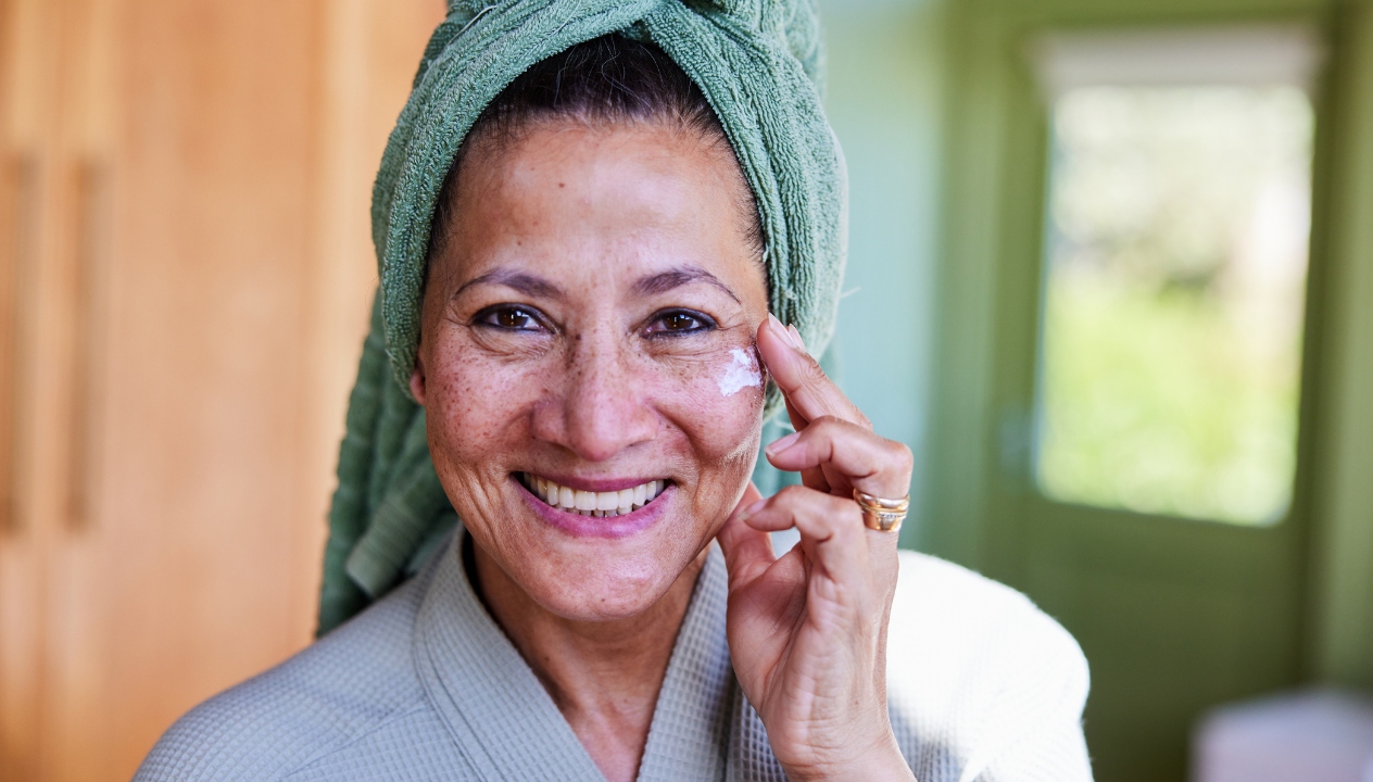 Smiling mature woman in a bathrobe applying moisturizer to her cheek