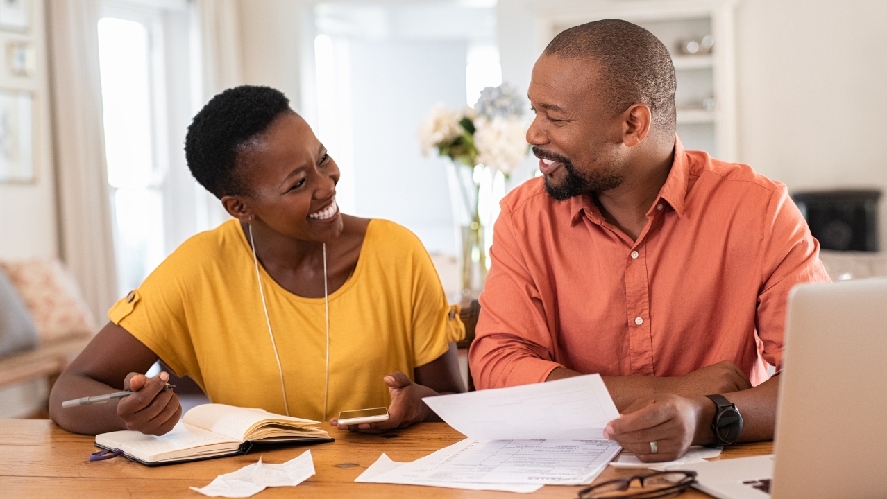 Couple smiling at each other while sitting at a table looking at documents.