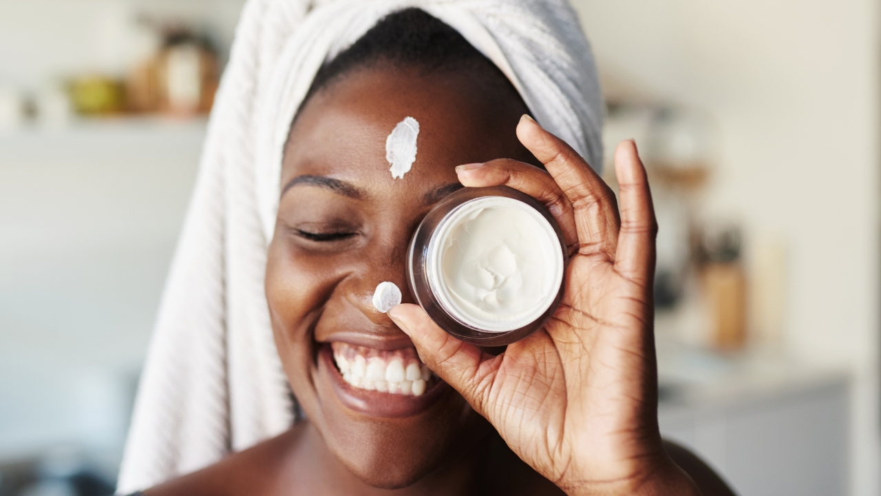 Person smiling while holding up a small jar of face cream with their hair in a towel.