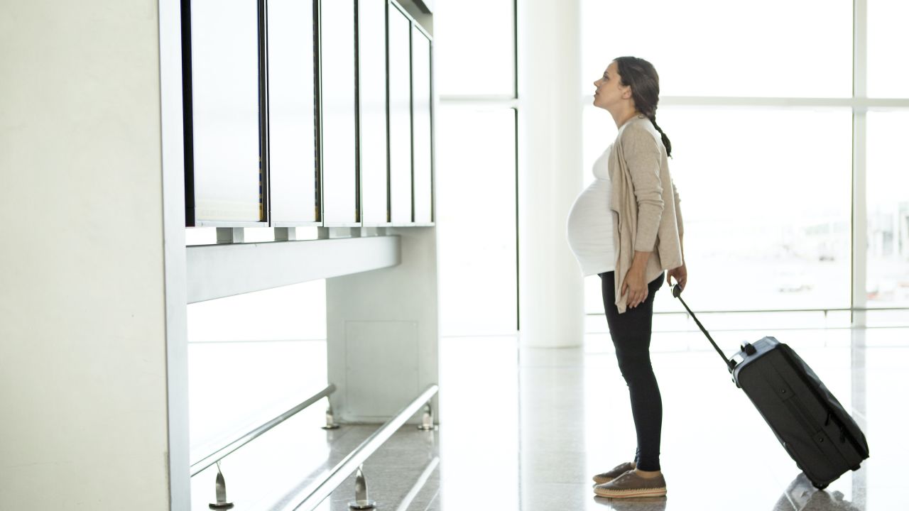 Pregnant person with a suitcase looking at an airport departure board.